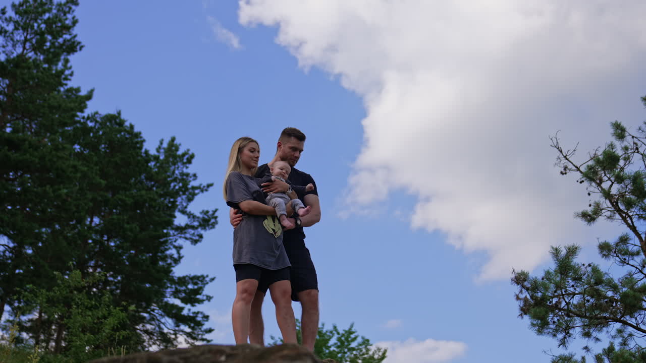 Caucasian couple stand embracing on the rock. Family of three with newborn baby in nature. Low angle view.