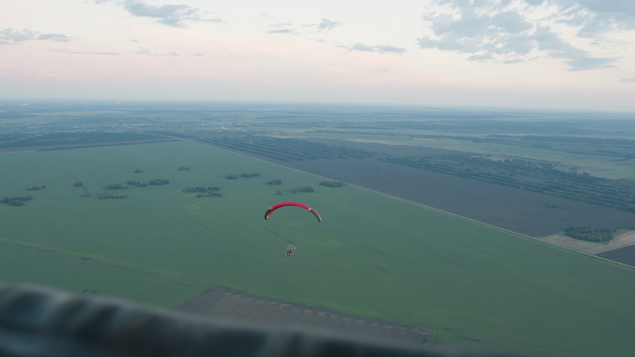 Overhead view of pilot flying powered paraglider over patchwork fields, captured from companion craft with framing bar in foreground against pastel evening sky conveying serene aerial motion