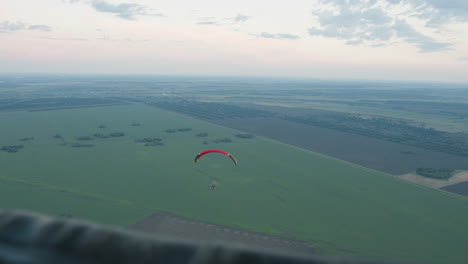 overhead view of pilot flying powered paraglider over patchwork fields, captured from companion craft with framing bar in foreground against pastel evening sky conveying serene aerial motion