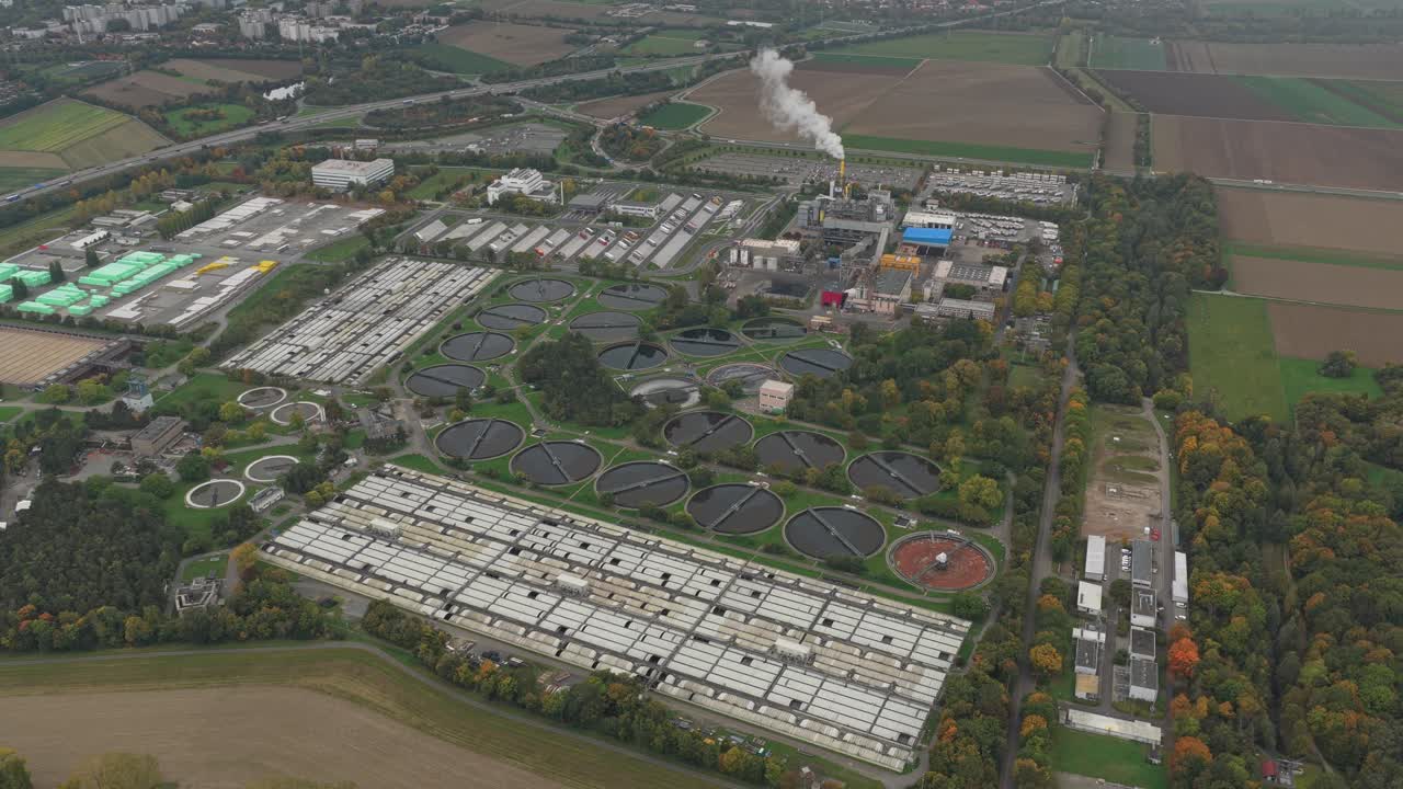 Wide aerial shot of a sewage treatment facility with circular settling tanks and aeration basins, surrounded by countryside and industry