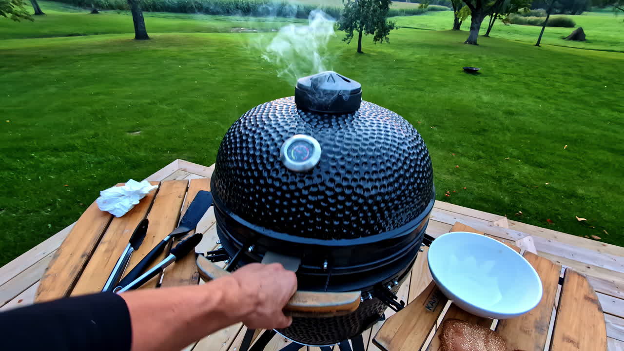 Grill smoking on a wooden deck in a lush green backyard with barbecue tools laid out