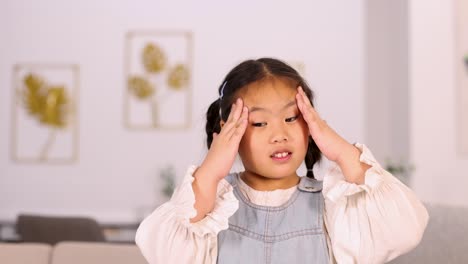 an asian child touches her temples with both hands, showing a headache gesture in a softly lit, modern living room with neutral decor