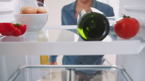 young woman reaching inside refrigerator and taking out bottle of wine and ready meal