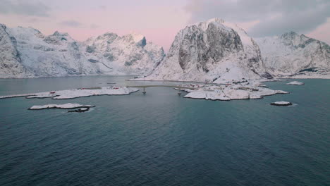 Moskenesøya-snowy-mountain-skyline-low-aerial-view-orbiting-idyllic-Reine-Norway-fishing-village