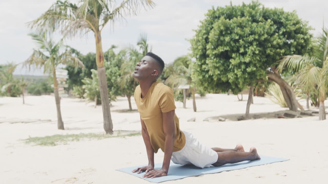 hombre afroamericano en forma haciendo yoga, estirándose en la alfombra en la playa, cámara lenta