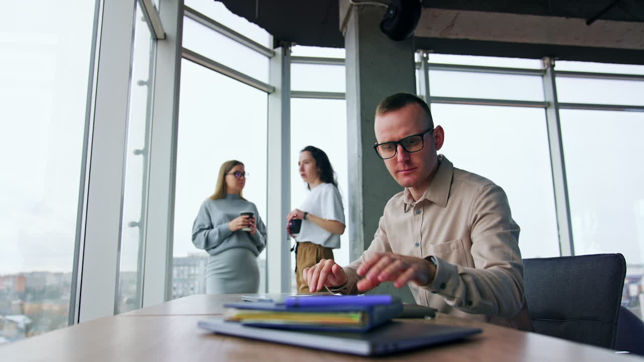 Puzzled and nervous male employee closes laptop and takes paper notebook to search the information. Man is happily rejoicing when founds it. Female colleagues gossiping at backdrop.