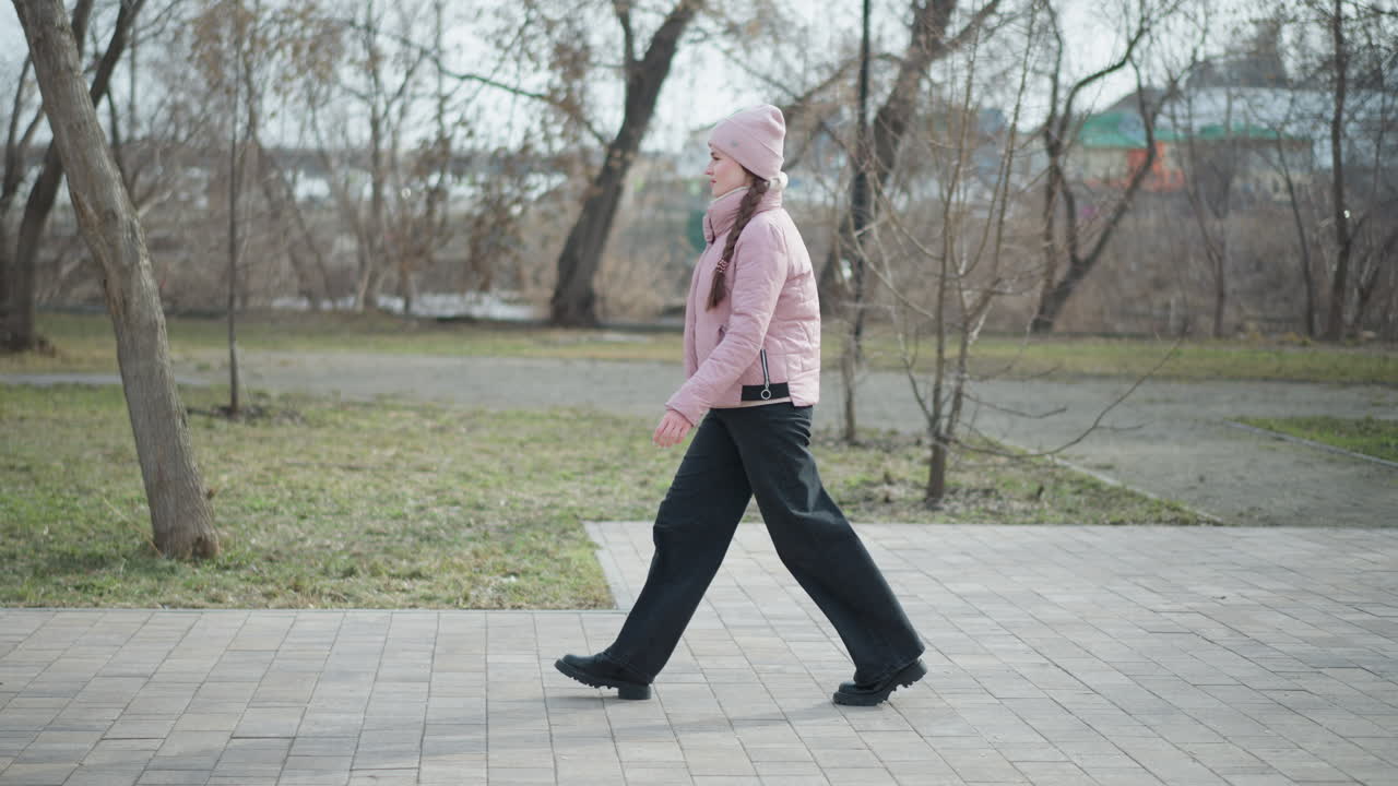 Side view of young woman wearing pink jacket and beanie walking on paved park path during cold winter day, surrounded by bare trees and muted nature, capturing movement, mood, and seasonal atmosphere