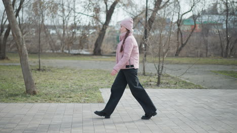 side view of young woman wearing pink jacket and beanie walking on paved park path during cold winter day, surrounded by bare trees and muted nature, capturing movement, mood, and seasonal atmosphere