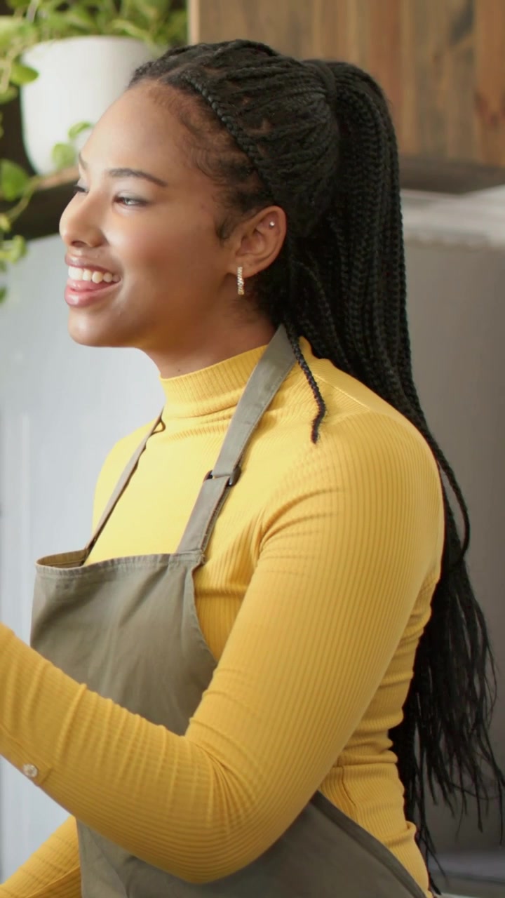 Vertical video: Smiling woman wearing apron lifting gaze, turning and reaching, serving in kitchen