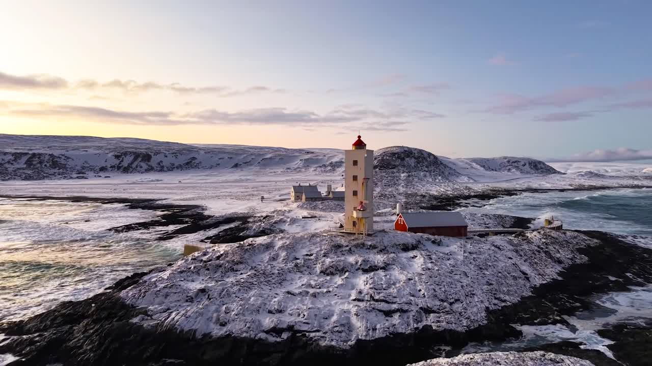 Aerial drone footage of Kjølnes or Kjolnes Lighthouse in Norway during a sunny day. The lighthouse is white and stands on snow covered shoreline between arctic ocean sea water that is wavy and foamy.