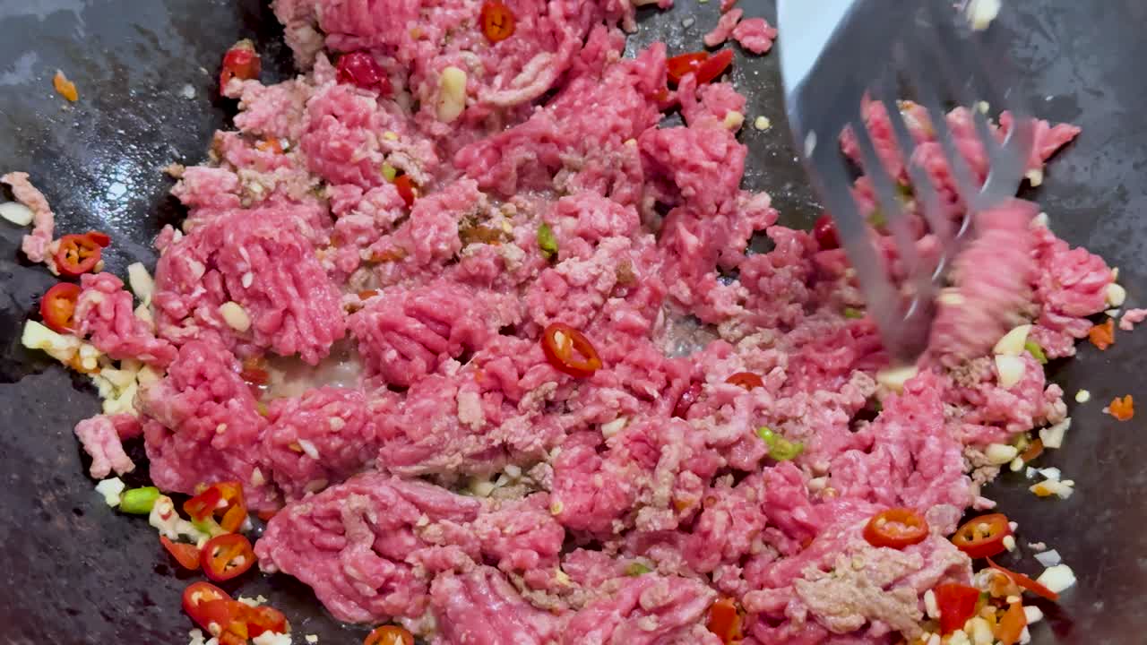 Raw minced meat is stir-fried with chopped red chilies and garlic in a wok under bright lighting, using a spatula for mixing