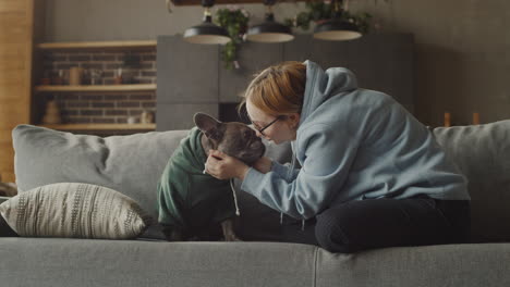 red haired woman puts a hood on her bulldog dog head and caresses her while sitting on the couch in the living room at home