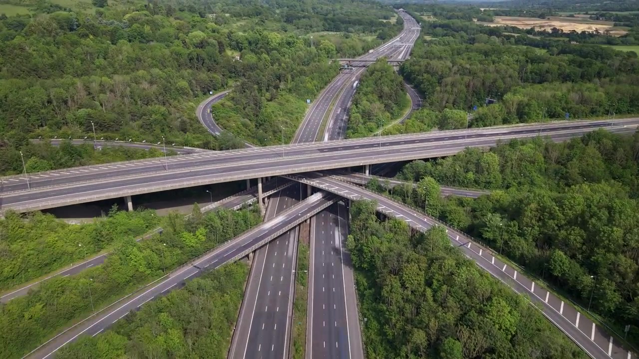 Aerial hyperlapse capturing steady traffic flow across a complex multi-level highway interchange. Vehicles streak through the intricate road network surrounded by lush green forest during the day.