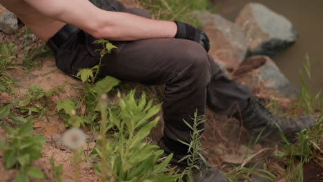 close-up view of warrior's booted legs stepping down uneven riverbank slope, pressing into loose soil and dandelion-covered grass during cloudy daylight, surrounded by natural greenery and rough terrain