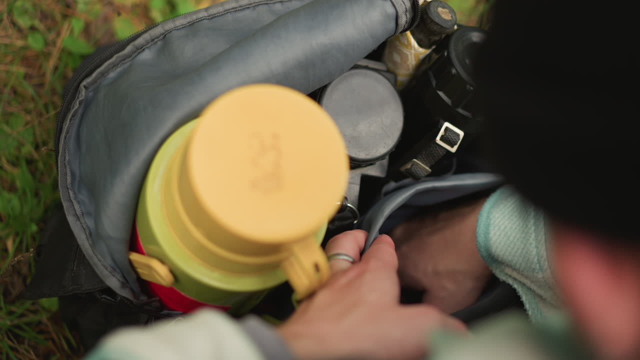 Escursionista caucasico che ripone gli ultimi oggetti nello zaino, vista dall'alto che mostra un thermos e una macchina fotografica tra l'attrezzatura nel campeggio erboso, mani che liscia il tessuto e fissa le cinghie, silenziosa attesa