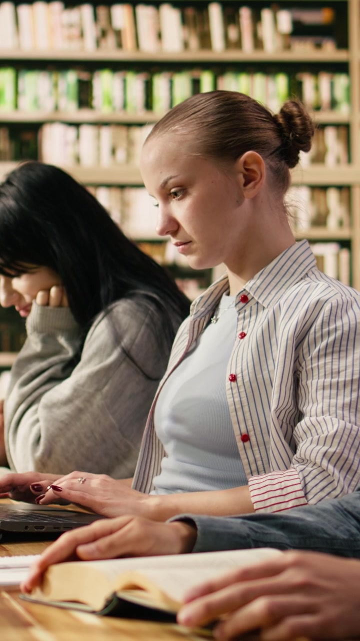Video verticale Studente che digita al portatile e prende appunti da un libro aperto a una scrivania in biblioteca