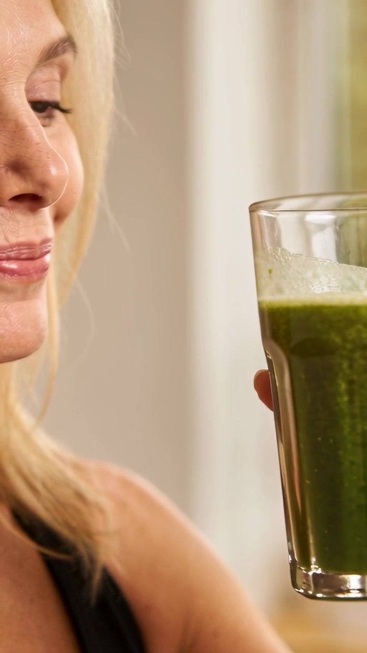 Dikey video: Mutfakta siyah atlet giyen bir kadın, bardağı kaldırıp yeşil smoothie tadıyor.