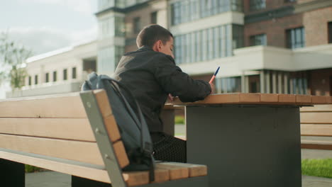 Behind view of boy sitting on bench outdoors writing with pen at wooden table near school building, backpack resting beside him, capturing focused learning moment in natural daylight