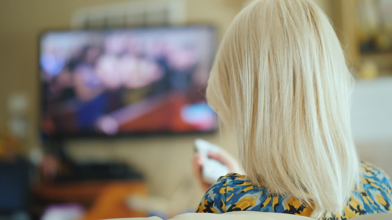 한 여성이 거실 뒷모습에서 tv를 보고 있으며 리모콘 4k 비디오를 들고 있습니다.