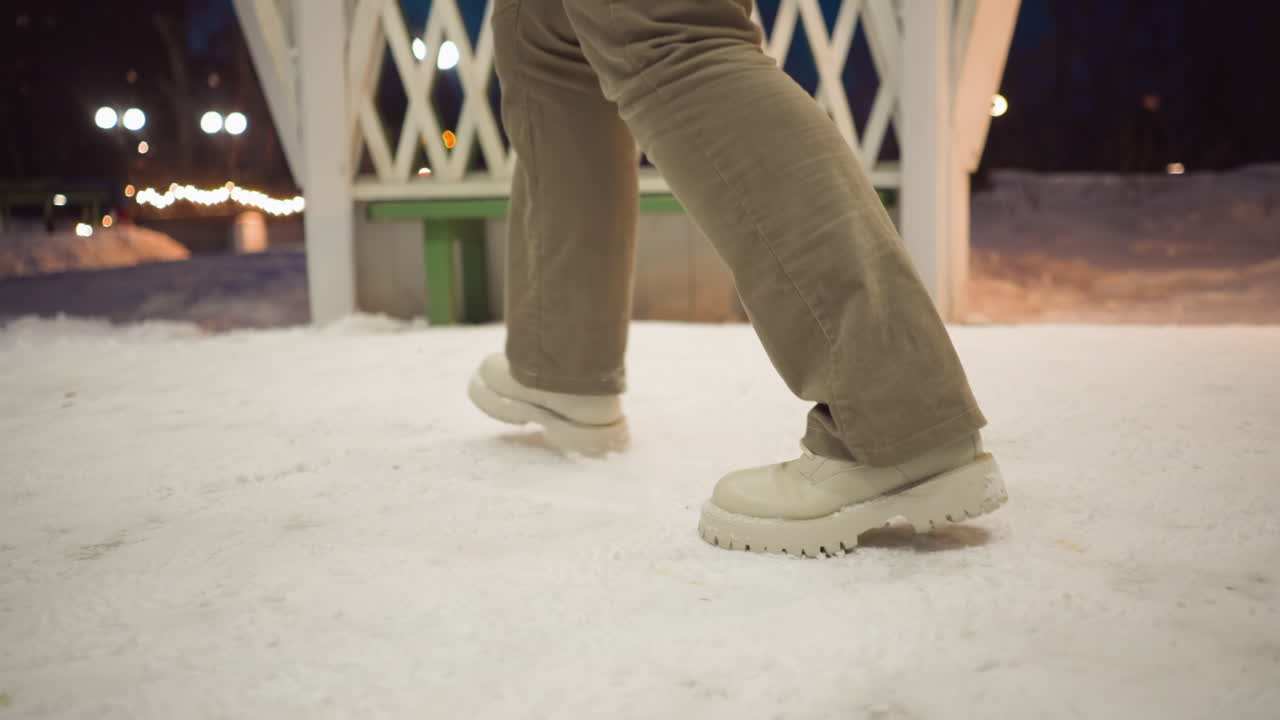 Girl wearing beige boots and khaki pants swirls legs inside light painted gazebo with snow beneath feet at dusk surrounded by glowing fairy lights and cold winter air conveying playful movement
