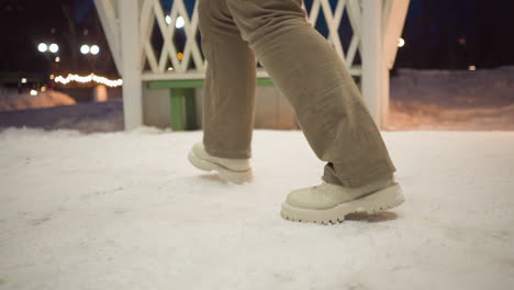 girl wearing beige boots and khaki pants swirls legs inside light painted gazebo with snow beneath feet at dusk surrounded by glowing fairy lights and cold winter air conveying playful movement