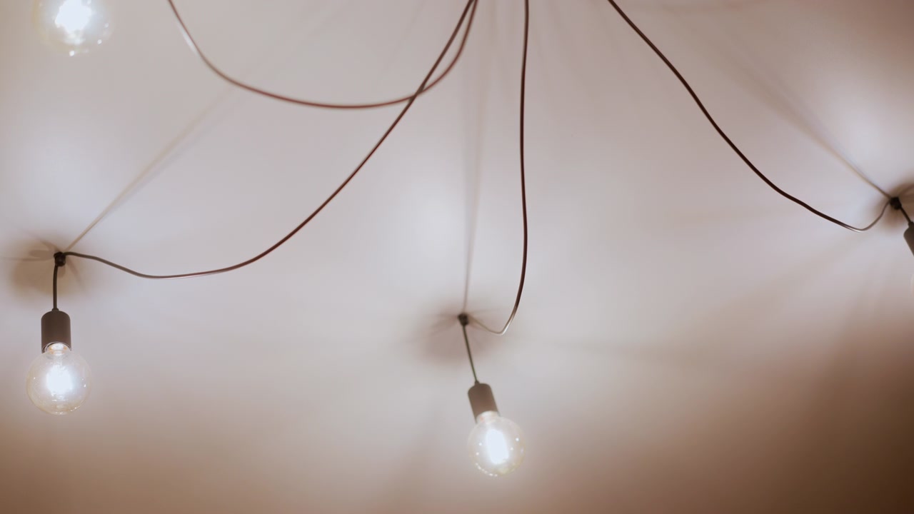 Primo piano di più lampadine a vista appese al soffitto con fili visibili che brillano delicatamente e creano un'illuminazione ambientale calda. Le lampadine sono sospese a diverse altezze su sfondo bianco semplice.