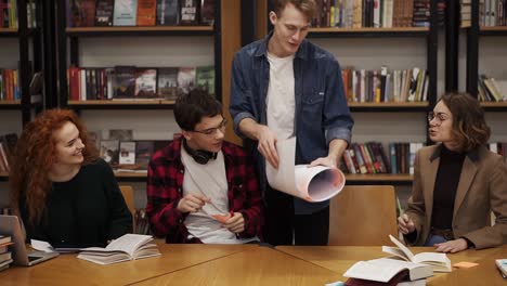 vista frontal de jóvenes estudiantes europeos, equipo de investigación de marketing haciendo una lluvia de ideas trabajando en un plan de negocios de inicio, estadísticas usando tecnología de tabletas portátiles. el tipo rubio trae el plan en papel y lo pone sobre la mesa