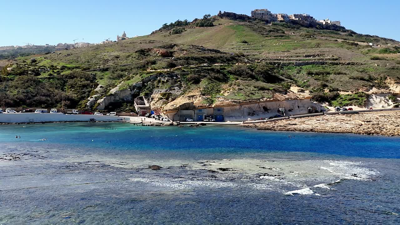 Vista panoramica mozzafiato della baia di Marsalforn, Gozo, Malta, sotto il sole estivo, con le vivaci acque turchesi del Mediterraneo incastonate tra le rustiche scogliere calcaree e l'architettura collinare in lontananza