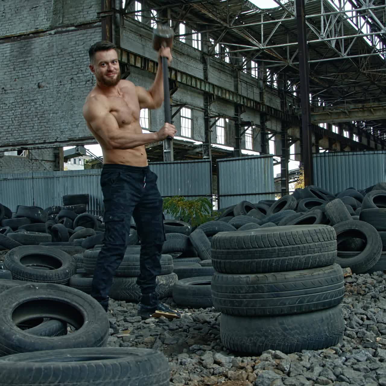 Sportsman with a heavy hammer in old plant. Sporty man trying to break pile of car tires with a metal hammer and smiles to camera. Workout in abandoned place.