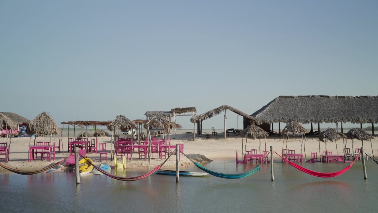 Toma panorámica estática de una playa tropical con coloridas hamacas sobre el agua, cabañas de palapa y muebles de exterior rosas sobre arena blanca
