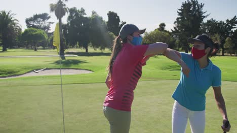 Two-caucasian-women-playing-golf-wearing-face-masks-greeting-each-other-with-elbows
