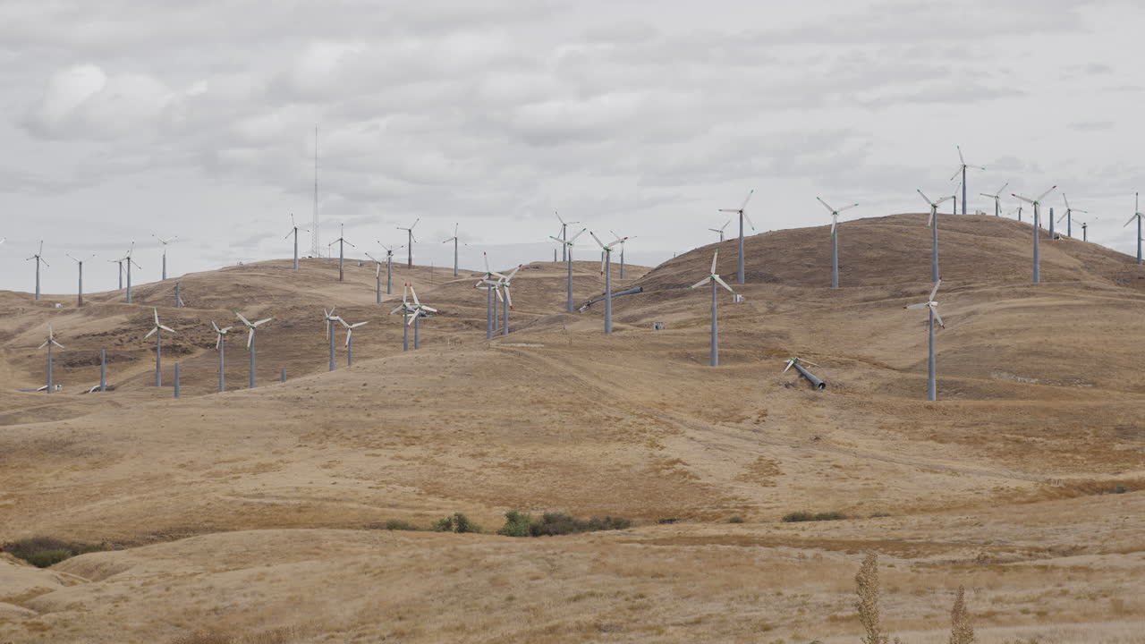 A large group of windmills not turning, some broken, on dry hill on cloudy day