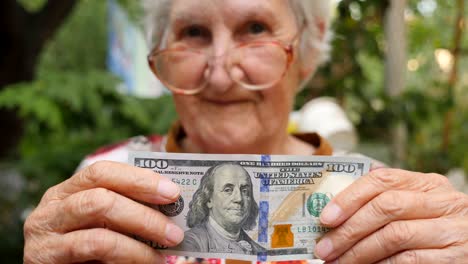 retrato de una anciana con gafas que muestra un billete de cien dólares en la cámara al aire libre. abuela feliz sosteniendo moneda extranjera afuera. concepto de dinero. cerrar la cámara lenta