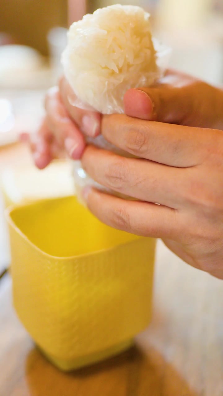 Manos desenvuelven arroz pegajoso del plástico en un recipiente de mimbre amarillo sobre una mesa de madera, con luz natural.
