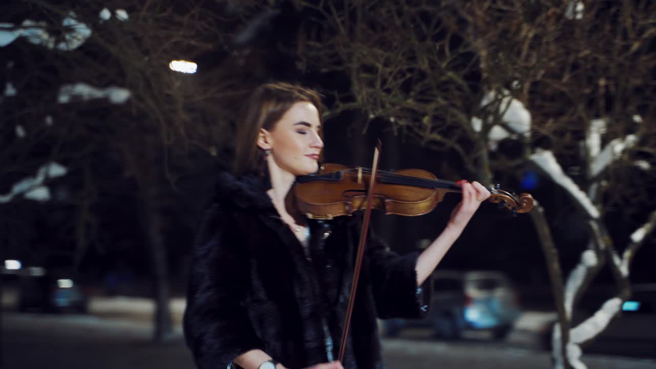 A beautiful girl with make-up and in a white dress is playing a violin composition in a winter park on the background of snow. Camera motion to left