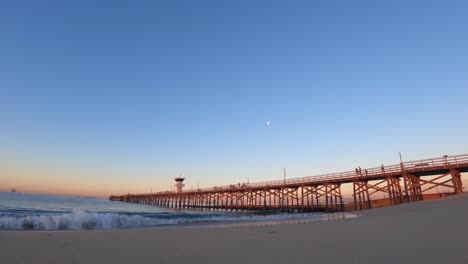 the moon setting over the beach with a long wooden pier at sunrise - seascape time lapse