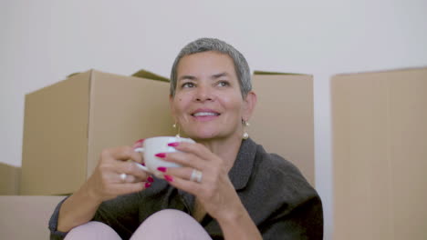 happy woman sitting on floor, drinking tea after moving to new house