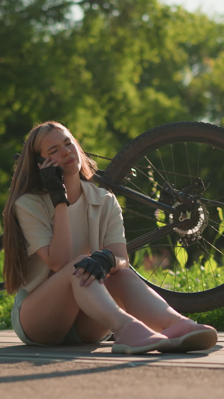 어린 여인이 거꾸로 자전거 근처에 앉아서 걱정스러운 표정으로 전화에 응답하고, 야외 환경에서 울창한 초록색과 나무로 둘러싸여 있습니다.