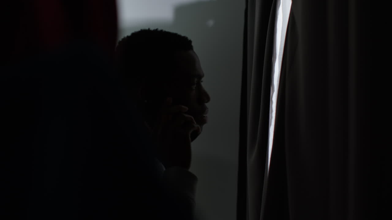 Young Man With Mental Health Issues Suffering With Paranoia Or Agoraphobia At Home Talking On Mobile Phone Looking Out Of Window