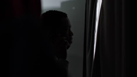 young man with mental health issues suffering with paranoia or agoraphobia at home talking on mobile phone looking out of window