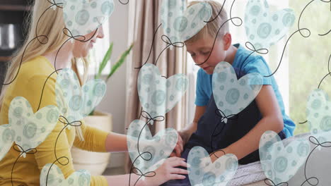 animación de globos de corazón sobre una madre caucásica feliz atando un zapato a su hijo en casa