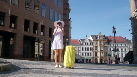 vertical video a slim young girl in a cute white dress and hat stands with a yellow suitcase in the old city. a french woman tourist takes a photo of the architecture of the old beautiful city