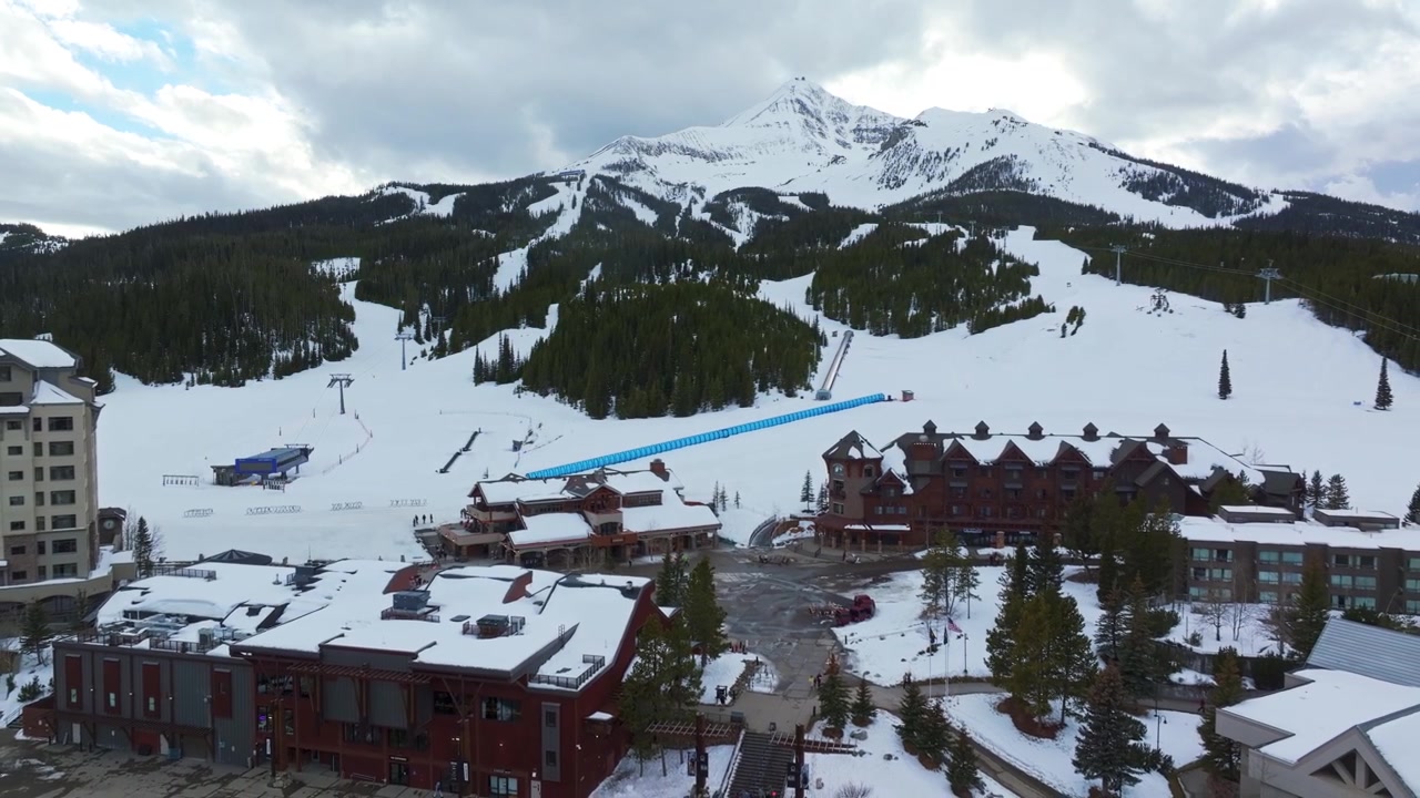 Loitontava laaja ilmakuva dronella Montanan vuoristokeskuksen talvimaisemasta, taustalla kohoaa Lone Peak, jota ympäröivät mäntymetsät, kodit ja lumipeitteinen maasto.