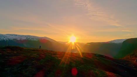 a lone person stands on a mountain ridge looking over a fjord in the scandinavian landscape during a golden sunset - dramatic aerial flyover