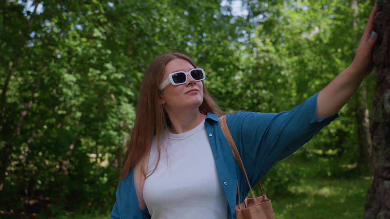 Young girl in white top and sunglasses walks through bright green park toward tall tree trunk, touching rough bark with admiration while sunlight filters through lush leaves in gentle summer breeze