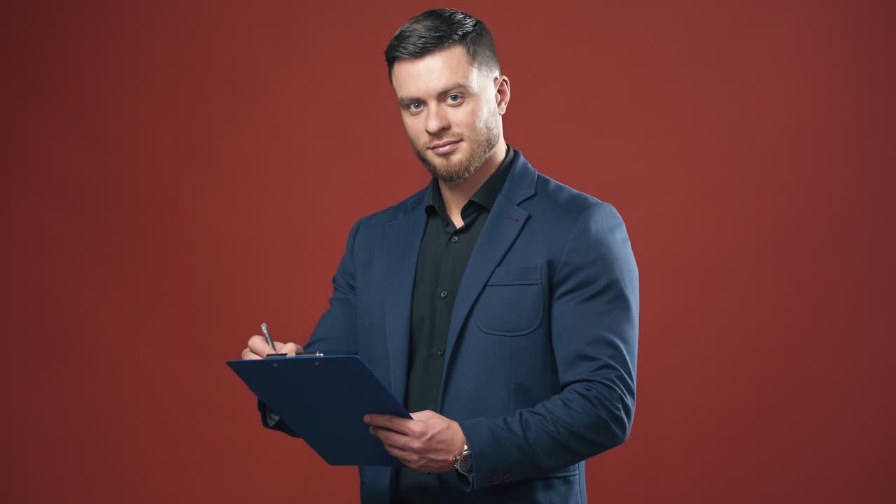 Businessman on red studio background. Portrait of a handsome man in dark suit. Successful man holding a folder and writes on it indoors.