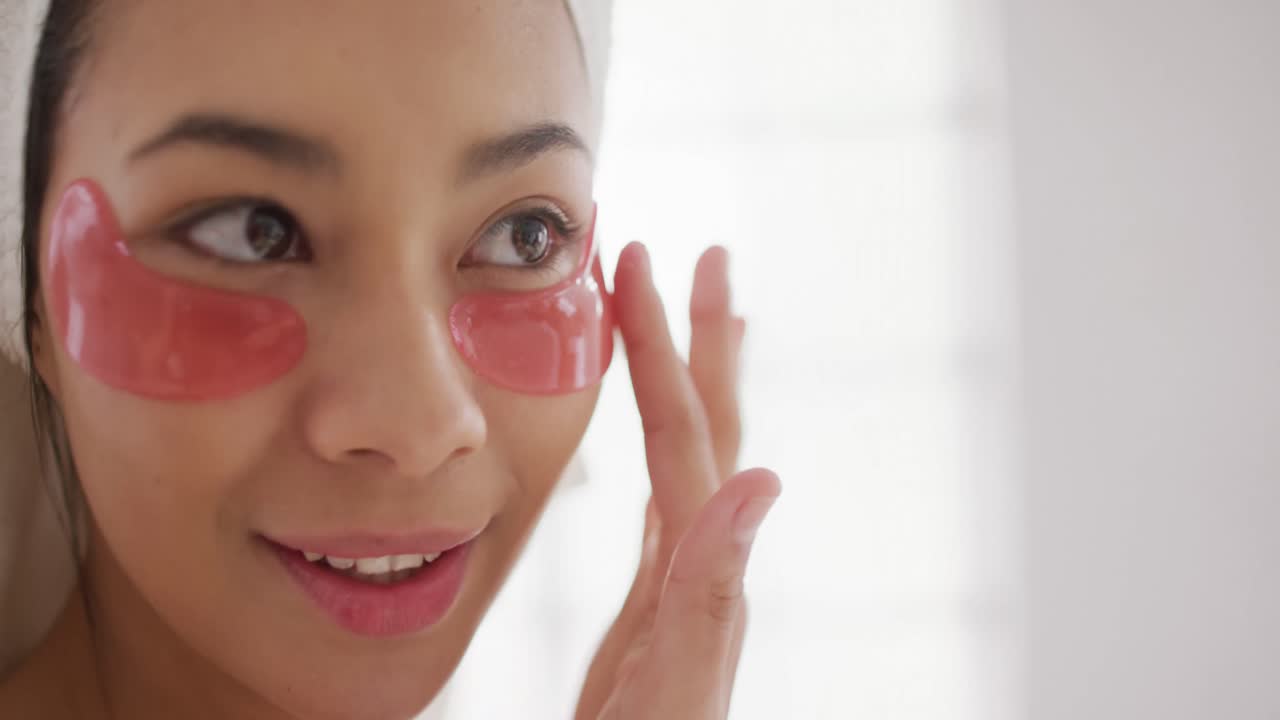 Video of portrait of smiling biracial woman with towel on hair applying eye mask in bathroom