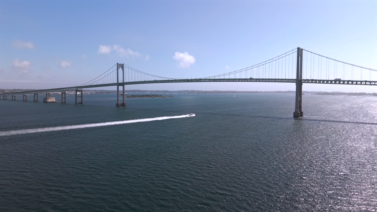 An aerial view from the Taylor Point Restoration Association look out on a sunny day with blue skies. The Claiborne Pell-Newport Bridge is in view as a boat speeds. The camera truck left