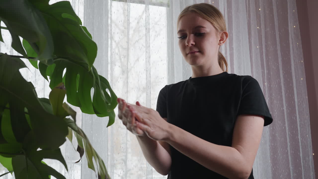 Young light skin woman wearing black shirt gently rubbing cream in hand while standing beside large green indoor plant with soft daylight coming through sheer white curtain in cozy serene setting