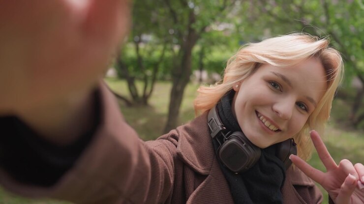 Donna scatta un selfie all'aperto facendo il segno di vittoria e sorridendo. La donna indossa cuffie attorno al collo con uno sfondo di parco verde con alberi e un cartello all'aperto